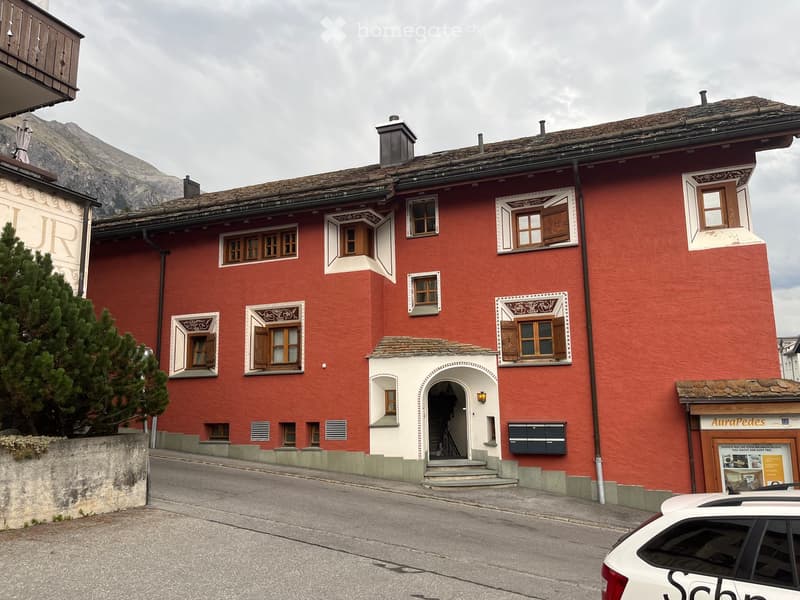 A three-story red building with white decorative window frames and shutters, featuring a central arched entrance and a small staircase leading up to it. The building is situated in a mountainous area with a small tree and a parked car visible in the foreg