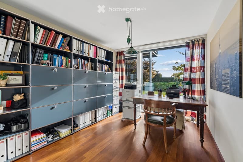 A well-organized office space with a large bookshelf filled with books and binders, a wooden desk with a chair, a lamp, and a printer. Large windows with curtains offer a view of the outdoors.