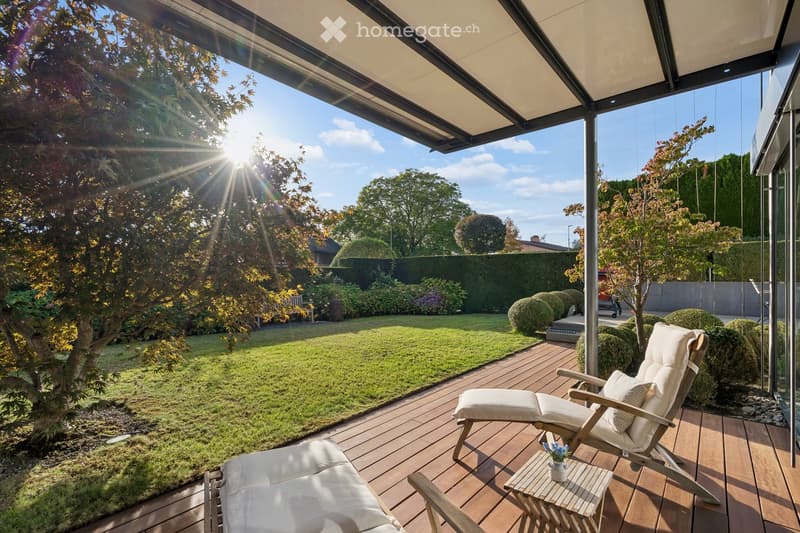 A sunny outdoor patio with wooden decking, lounge chairs, a small table with a flower pot, and a well-maintained garden with grass, trees, and shrubs.