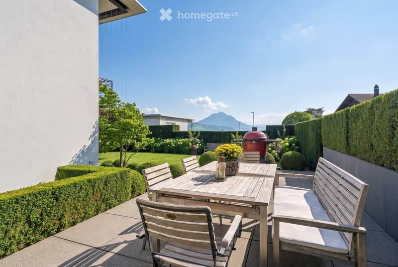 A well-maintained garden with a wooden patio area, featuring a dining table, chairs, a bench, and a barbecue grill. The garden is surrounded by neatly trimmed hedges and trees, with a scenic mountain view in the background.