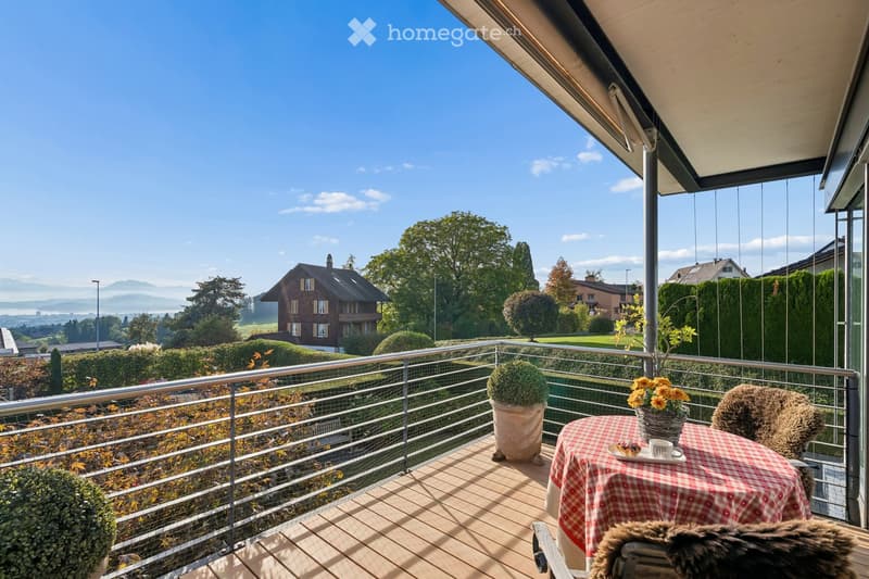 Balcony with wooden decking, metal railing, table with checkered cloth, flower pot, and cup, overlooking a scenic landscape with houses and mountains.