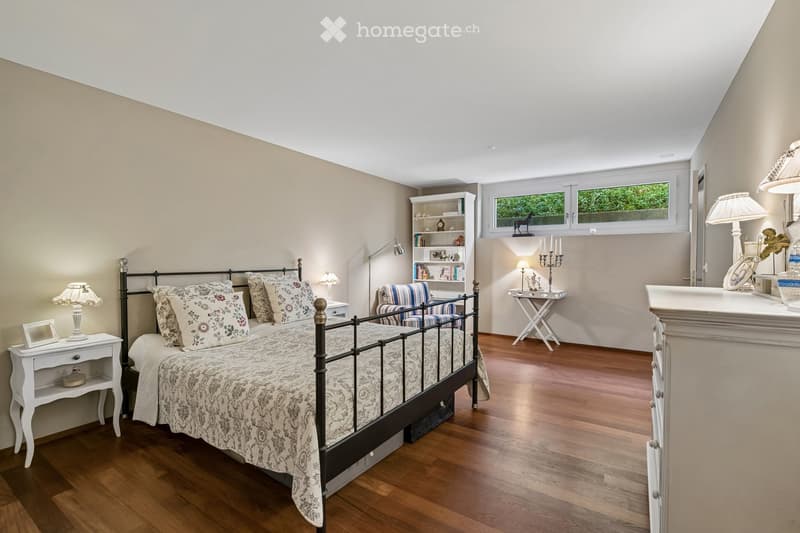 Cozy bedroom with wooden flooring, a black metal bed with floral bedding, nightstands with lamps, a small armchair, a bookcase, a small table with a candle holder, and a dresser with a mirror.