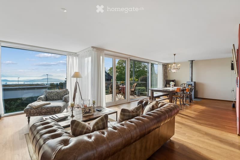 Bright living room with large windows offering a scenic view, featuring a leather sofa, coffee table, dining area with chairs and table, fireplace, chandelier, and floor lamp.