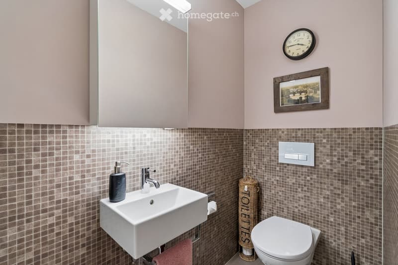 Modern bathroom with a white sink, chrome faucet, toilet, mirror, clock, picture frame, soap dispenser, wicker basket, and a red towel.