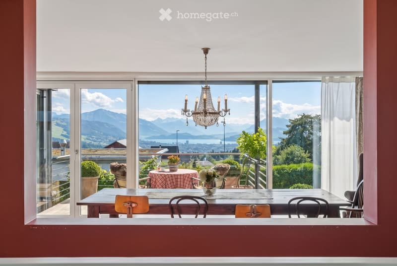 A well-lit room with a large wooden dining table, chairs, a chandelier, and a vase on the table. Large windows offer a scenic mountain view.