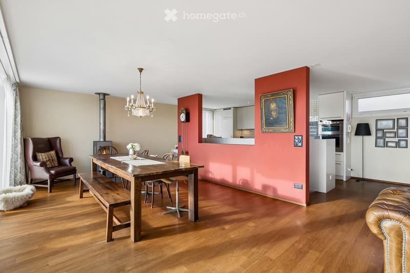 Open-plan living and dining area with wooden flooring, a wooden dining table with bench and chairs, leather armchairs, a chandelier, a fireplace, and a modern kitchen area with white cabinets and stainless steel appliances.