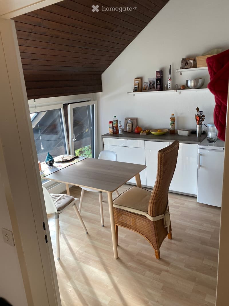 A modern kitchen with a wooden dining table, two chairs, white cabinets, and a refrigerator. The room features a slanted wooden ceiling and a large sliding glass door leading to a balcony.
