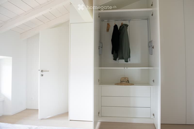 A minimalist, unfurnished room with a built-in white closet featuring a hanging rod, shelves, and drawers. The room has light-colored walls, a wooden floor, and a simple door.
