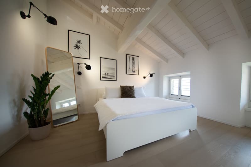 Bright, airy bedroom with white walls, wooden floor, slanted wooden ceiling, large bed with white linens, decorative pillows, wall-mounted lamp, large mirror, potted plant, and framed artwork.