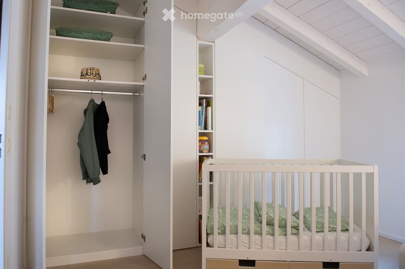 A cozy, well-lit bedroom with a white crib, green pillows, and a matching blanket. A built-in wardrobe with open shelves and a small bookshelf is visible, containing books and decorative items.