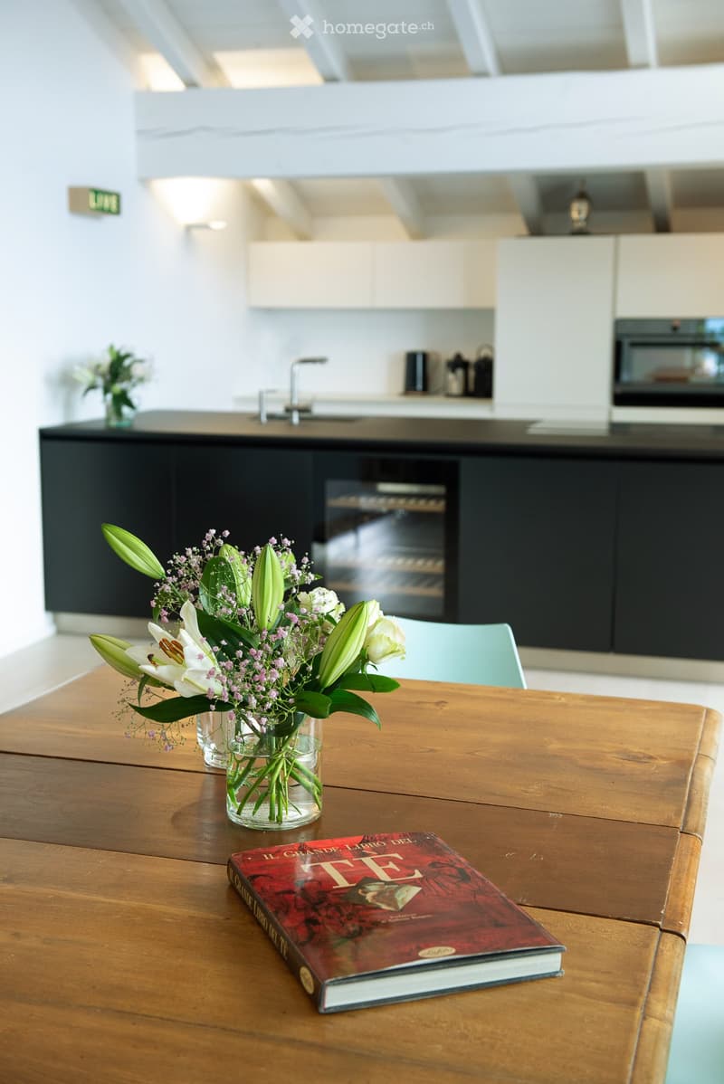 Modern kitchen with a wooden dining table, vase with flowers, and a book on the table. The kitchen features dark cabinets and stainless steel appliances.