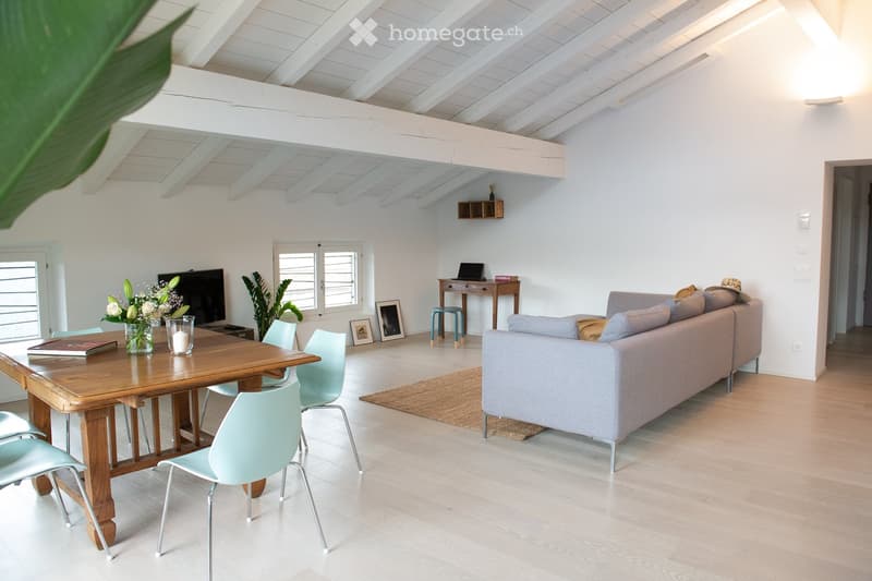 Bright living room with white walls, wooden floor, and high ceiling. Features a grey sofa, wooden dining table with chairs, a small desk with a laptop, and decorative items including a vase with flowers and a candle.
