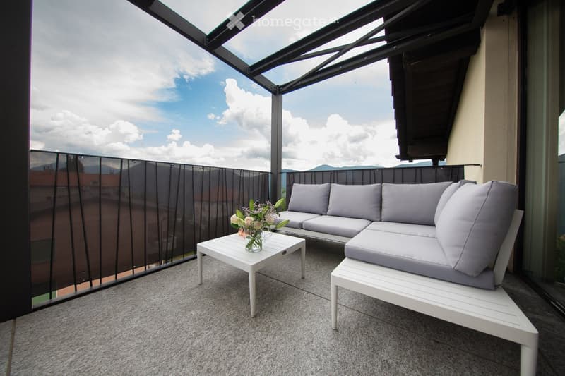Balcony with a modern white sectional sofa, a small white coffee table with a flower vase, and a view of the sky and distant buildings.