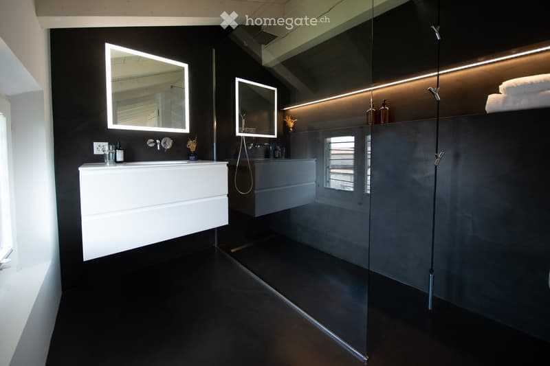 Modern bathroom with dark walls, white sink, illuminated mirrors, glass shower enclosure, and sleek fixtures.