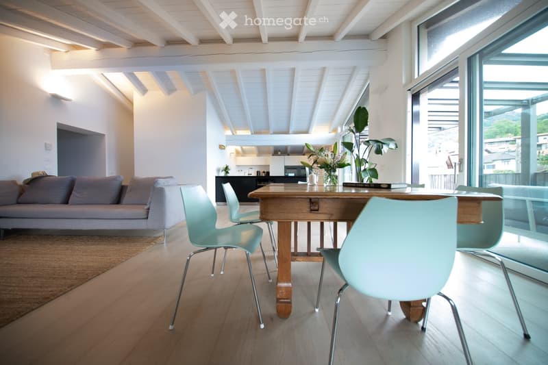 Open-plan living and dining area with a grey sofa, wooden dining table, modern chairs, and a visible kitchen area with cabinets and appliances.