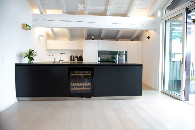 Modern kitchen with white cabinets, black lower cabinets, a wine cooler, and a sleek sink area. The space features a light wooden floor and an open, airy design with a visible living area above.
