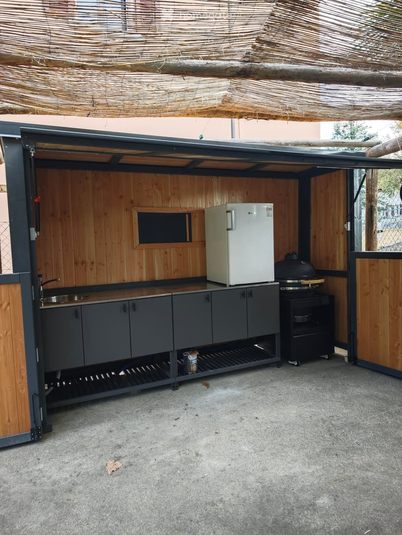 Compact outdoor kitchen area with wooden walls, stainless steel sink, refrigerator, cabinets, grill, and oven, all under a thatched roof.