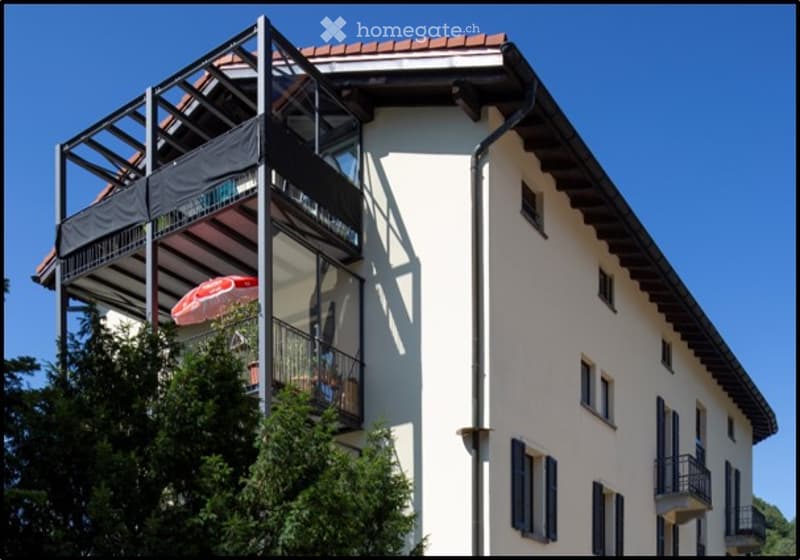 A modern multi-story residential building with balconies, large windows, and a red-tiled roof under a clear blue sky.
