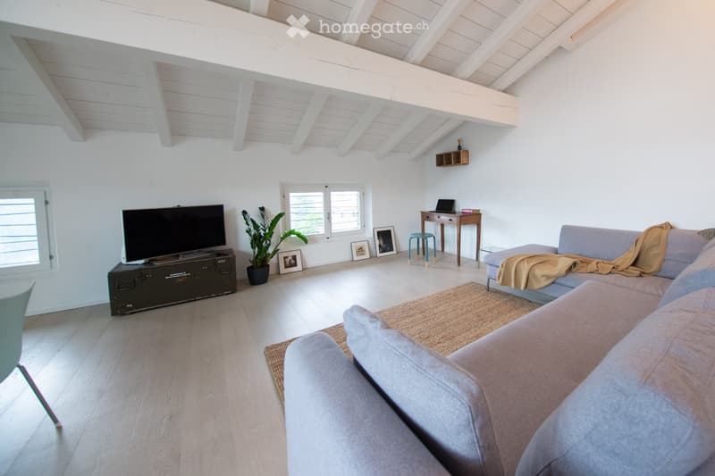Bright living room with white walls, wooden floor, exposed wooden beams, large windows, sofa, coffee table, laptop, stool, small table, television, plant, and picture frames.