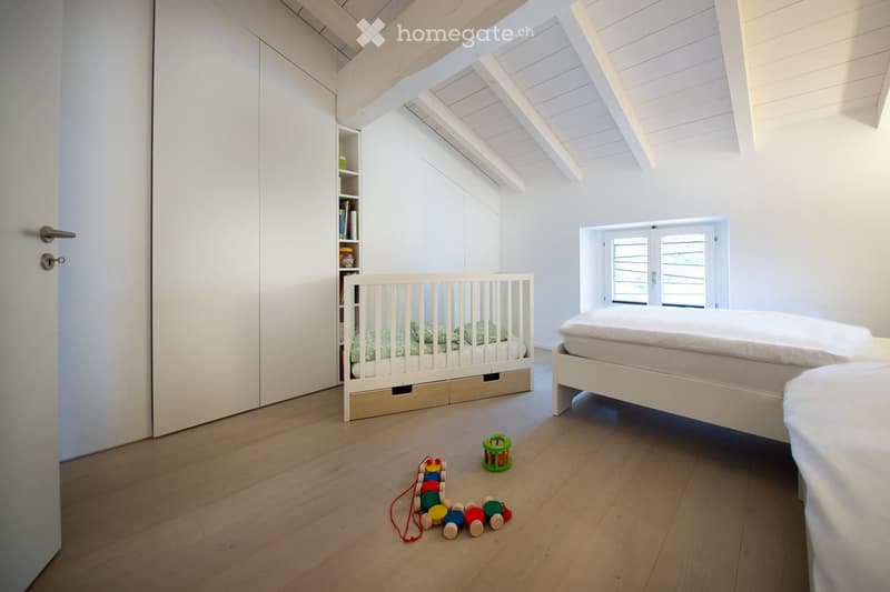 Bright, airy bedroom with wooden flooring, white walls, and a slanted ceiling. Features a double bed, a crib, a built-in wardrobe, a small shelf, and scattered toys on the floor.