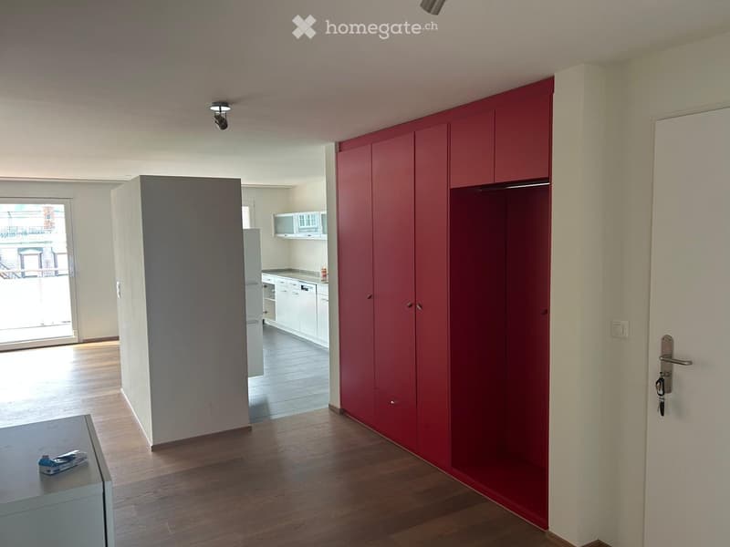 A modern, unfurnished room with wooden flooring, a large red wardrobe, a small table with a few items, and an open kitchen area with white cabinets and a dishwasher.