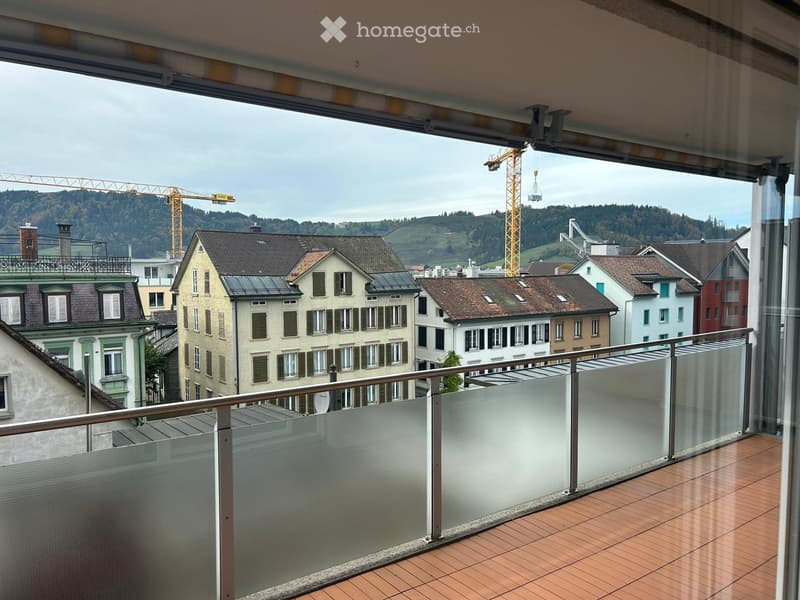 Balcony with glass railing, tiled floor, overlooking a cityscape with buildings and hills.