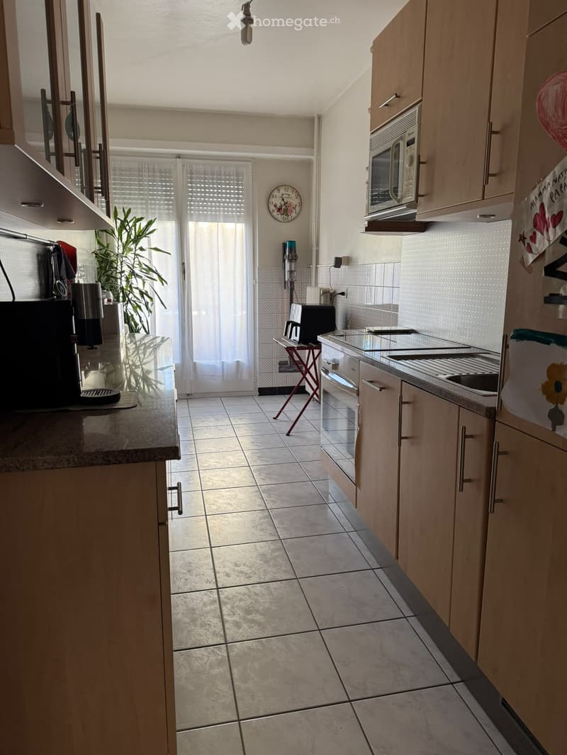 Bright kitchen with light wooden cabinets, tiled floor, microwave, oven, sink, coffee maker, and a small stool near the door.