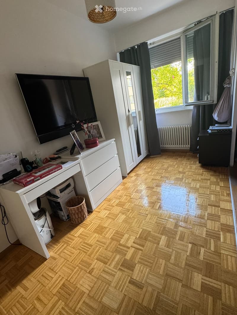 Well-lit room with herringbone wooden flooring, large window with dark curtains, white desk with chair, television mounted on wall, wardrobe with mirror, and various decorative items including picture frames and a wicker basket.