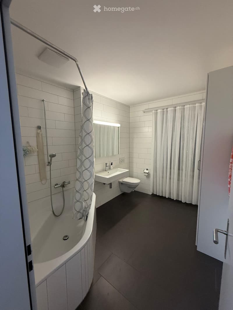 Modern bathroom with a bathtub, shower curtain, sink, mirror, and toilet. The walls are tiled, and the floor is dark.