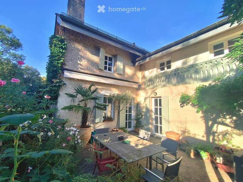 Courtyard with a wooden table and chairs, surrounded by potted plants and flowers, with a building featuring shutters and a balcony in the background.