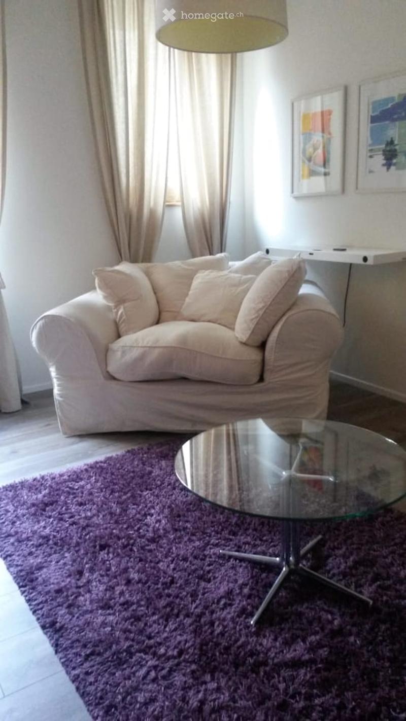 A cozy living room with a light-colored sofa, a glass coffee table, a purple shag rug, and a hanging lamp. The room features large windows with curtains and framed artwork on the wall.