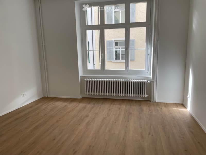 Empty room with wooden flooring, white walls, and a large window with a radiator below it.