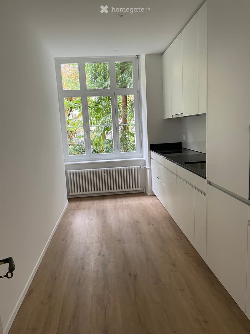 Empty kitchen with light wooden flooring, white cabinets, and a large window showing greenery outside. No furniture or appliances are present.