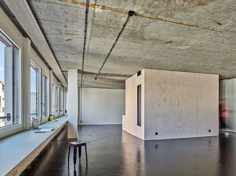 Empty room with exposed concrete ceiling, large windows, dark polished floor, and a small wooden stool near the window.