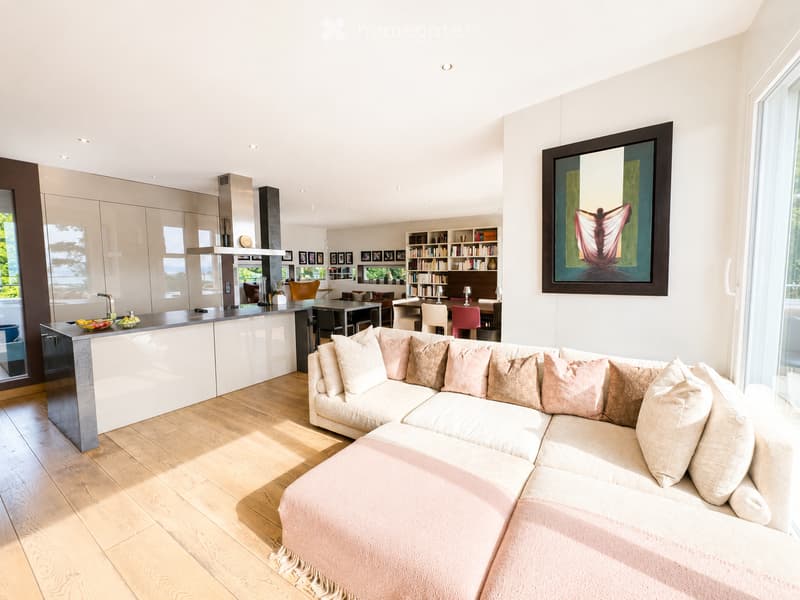 Modern living and dining area with a large sectional sofa, coffee table, kitchen island, bookshelf, dining table, and chairs. The space features light wooden flooring and large windows with a view.