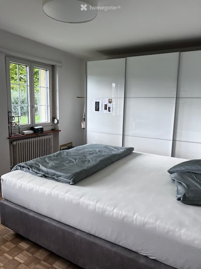 Modern bedroom with a large bed, grey bedding, white wardrobe, nightstand with a lamp, radiator, and picture frames on the wall.