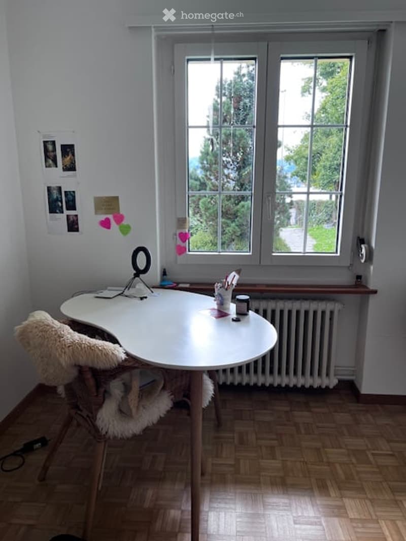 A modern, well-lit room with a large window, a white desk, a comfortable chair, and various small items on the desk.