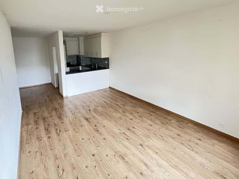 Empty room with light wooden flooring, white walls, and a small kitchen area visible in the background.