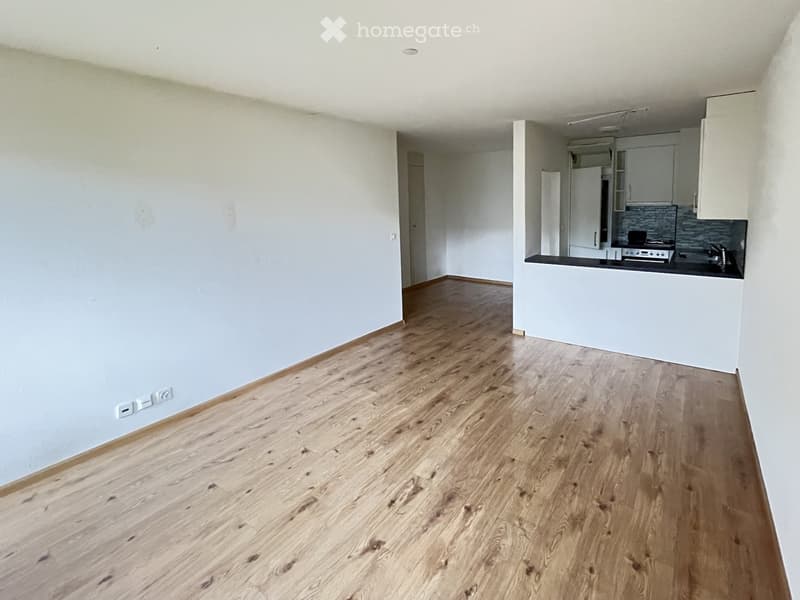 Empty room with light wooden flooring and white walls, featuring a small kitchen area with cabinets and appliances.
