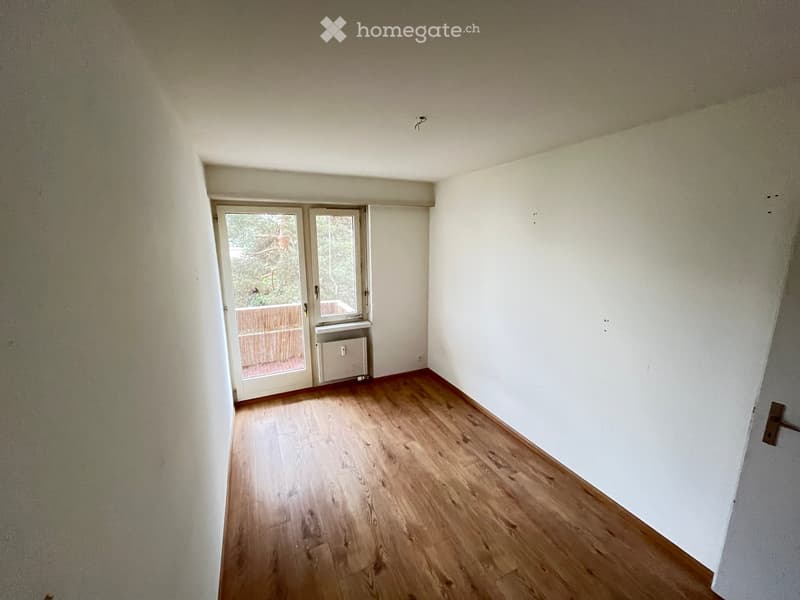 Empty room with wooden flooring, white walls, and a large window with a view of a wooden deck and greenery outside.