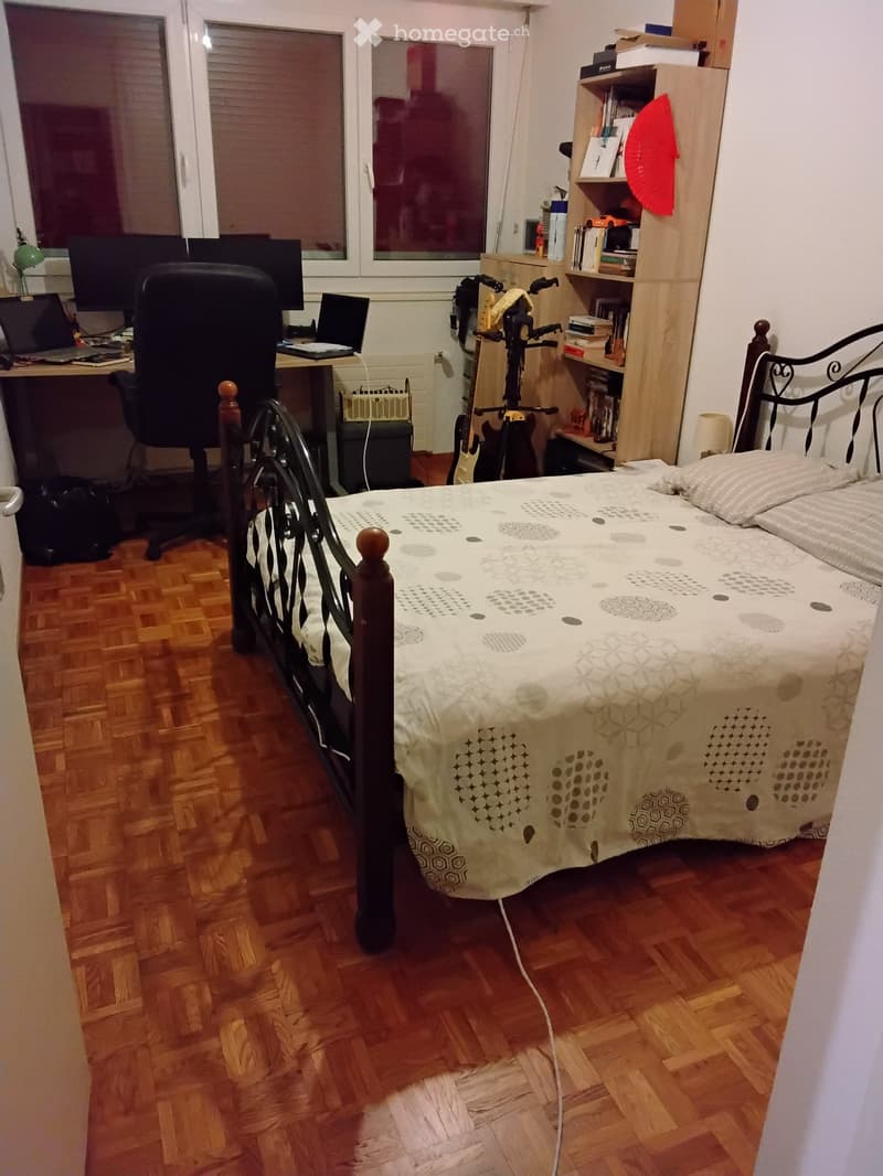 Bedroom with wooden flooring, a bed with patterned bedding, a desk with a laptop and chair, a guitar, and a bookshelf with books and decorative items.