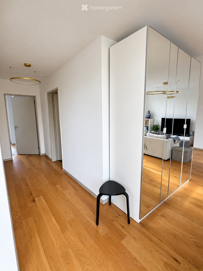 A modern hallway with light wooden flooring, white walls, and a large mirrored wall reflecting a living room area with a sofa, TV, and other furnishings. A single black stool is placed in the foreground.
