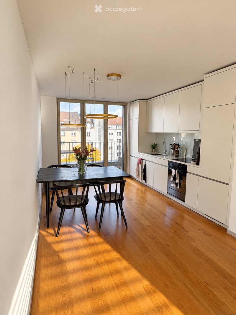 Modern kitchen and dining area with light wood flooring, white cabinets, a black dining table with four chairs, and a vase of flowers on the table. The kitchen features a sink, oven, and various utensils.