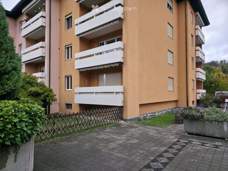 Multi-story residential building with multiple balconies, beige and pink facades, and a paved entrance area with greenery.