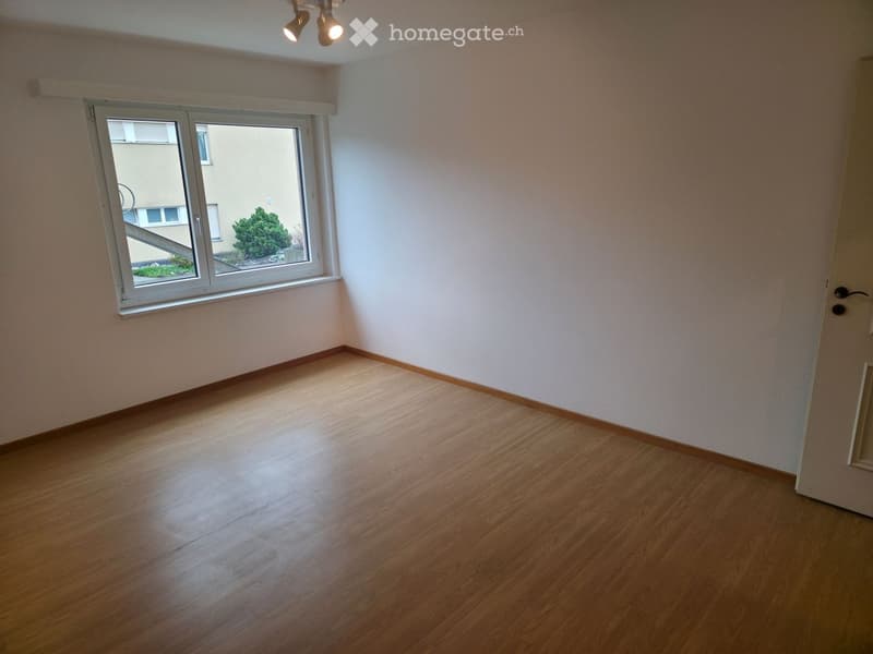 Empty room with wooden flooring, white walls, and a large window with a view of a small garden area.