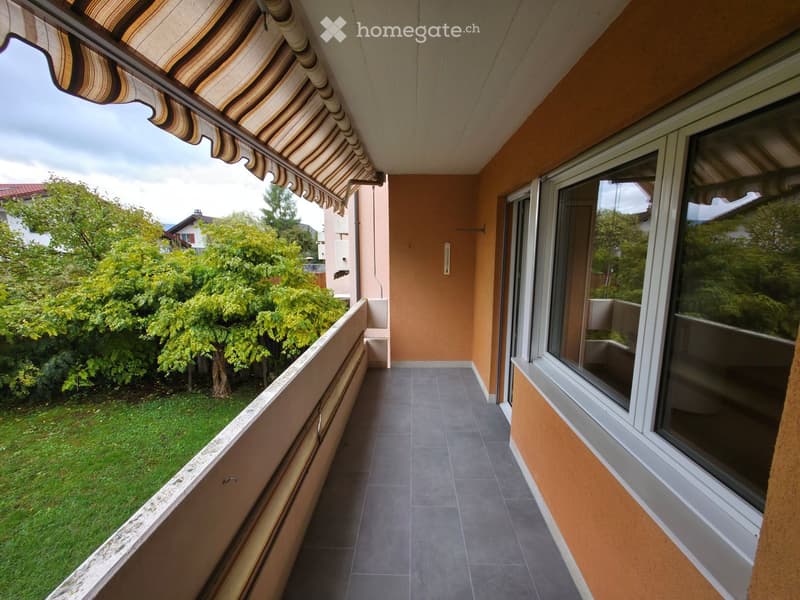 Balcony with tiled floor, striped awning, large windows, and view of a green garden with trees.