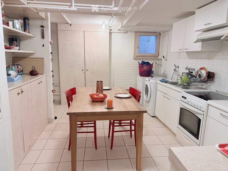 Bright kitchen with white cabinets, a wooden dining table with red chairs, an oven, washing machine, and various kitchen items on shelves and counters.