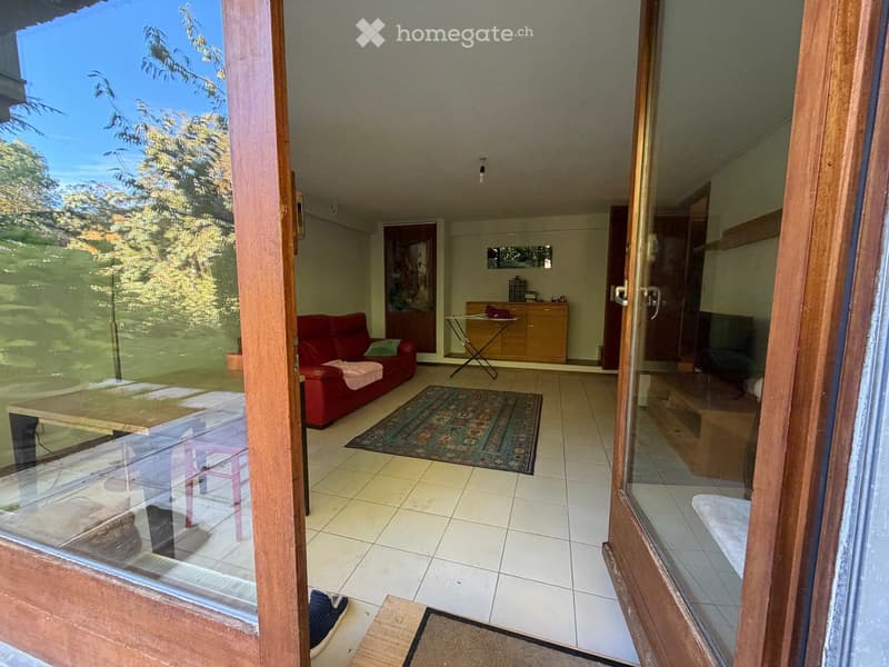 Bright living room with a red sofa, wooden table, patterned rug, and a view of a garden through large glass doors.