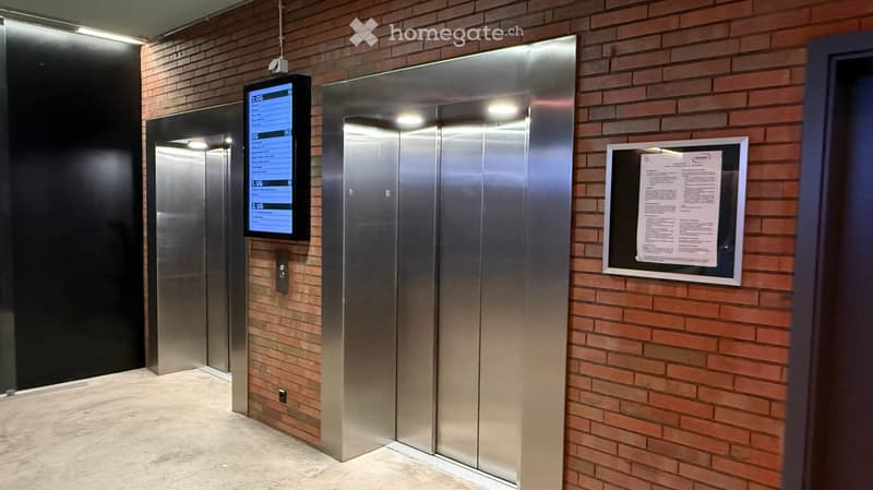 Modern hallway with brick walls, polished marble floor, and two elevator doors with digital displays showing floor numbers and status.