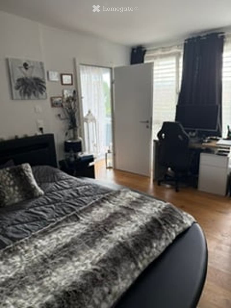 Cozy bedroom with a large bed, nightstand, desk with a computer, and a chair. The room features wooden flooring, a large window with dark curtains, and a picture frame on the wall.
